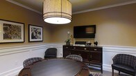 This single-floor space exudes a refined, club-like atmosphere with thoughtfully curated design elements throughout. The centerpiece of the room is a large, dark-finished round game table surrounded by plush plaid upholstered chairs, making it an ideal setting for social gatherings and leisure activities. A striking large-format drum pendant light with a warm woven shade hangs overhead, casting a welcoming glow across the space. Classic wainscoting lines the lower portion of the walls, complementing the rich, warm taupe tones of the upper walls and adding a layer of architectural sophistication. A substantial dark wood credenza serves as an anchor along one wall, topped with decorative accents including a delicate orchid arrangement and tasteful décor pieces. A wall-mounted flat-screen television above the credenza enhances the room's versatility for both entertainment and presentations. Framed black-and-white artwork adorns the walls, contributing to the space's polished and timeless aesthetic. Crown molding along the ceiling adds a finishing touch of elegance. Overall, the space balances functionality with upscale design, creating an inviting environment well-suited for socializing, gaming, or private gatherings.