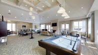 This sophisticated single-floor amenity space is designed to serve as a premier social and recreational hub. The interior immediately impresses with its soaring vaulted and coffered ceilings, adorned with a combination of stylish globe pendant chandeliers and industrial-style pendant lights that illuminate the space with a warm, inviting glow, complemented by recessed lighting throughout.

At the forefront is a full-size billiards table, positioned beneath statement pendant lights and set atop an intricately patterned carpet that anchors the recreational zone. Beyond the billiards area, the space transitions seamlessly into a comfortable lounge featuring plush seating arrangements centered around a classic white fireplace mantle, creating a cozy focal point for gatherings.

Flanking the lounge are dedicated dining and seating areas, offering ample space for social events or quiet relaxation. Tall, white-framed French doors and generously sized windows line the perimeter, flooding the interior with abundant natural light and providing views of the surrounding outdoor areas. Neutral tones of cream, taupe, and soft grey throughout the walls, furnishings, and architectural details contribute to the space's refined, elegant ambiance, making it a welcoming and versatile environment for a variety of activities.