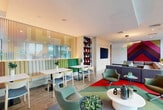 Located at 125 NE 32nd St in Miami's Upper Eastside neighborhood, this expansive single-floor amenity space offers a generously sized, thoughtfully designed communal environment ideal for socializing, lounging, and casual gatherings. The layout flows seamlessly from zone to zone, unified by warm wood-look tile flooring and crisp white walls throughout.

At the heart of the space is a sleek café-style bar counter with pendant lighting — a striking cascade of amber and tinted glass globes — paired with a built-in banquette and bar stools. Adjacent dining areas feature vibrant channeled banquette seating in tones of tan, sage, and emerald green, complemented by white bistro tables and mixed chairs. A large-scale cityscape mural adds artistic character to one dining nook.

The lounge area is anchored by generous sectional seating atop colorful area rugs, beneath modern circular pendant fixtures. A bold chevron-patterned accent wall in deep reds, purples, and magentas serves as a striking focal point. A billiards table adds a recreational element to the space.

Floor-to-ceiling glass curtain walls line the perimeter, flooding the interior with abundant natural light and offering unobstructed views of the lush outdoor terrace and surrounding landscape. The entry hallway features a curved glass facade, providing a bright, welcoming transition into the space.
