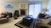 This single-floor space offers a warm and functional layout designed for comfortable everyday living. The living area serves as the focal point of the home, anchored by a classic brick fireplace with a wood mantle that adds a traditional, inviting character to the room. Natural light flows generously through a large sliding glass door dressed with vertical blinds, brightening the carpeted space and creating an open, airy feel. The living room is thoughtfully arranged with a plush sofa, a sleek black coffee table centered on a bold geometric area rug, and a leather recliner chair that adds both style and comfort. Wall art and decorative accents provide a personalized touch, while the neutral wall tones keep the atmosphere clean and understated. The overall ambiance is relaxed and homey, balancing practicality with comfort. The single-floor layout ensures easy accessibility and a seamless flow throughout the space, making it well-suited for a variety of lifestyles.