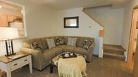 This two-story home offers a thoughtfully arranged living space designed for comfort and functionality. The main level features a cozy living room anchored by a large sectional sofa in warm, earthy tones, complemented by decorative throw pillows in floral and striped patterns. A wooden-framed mirror with ornate carved detailing adorns the wall, adding a touch of character to the neutral color palette. A low coffee table with a tray and decorative accents sits at the center, draped with a light knit throw that enhances the room's relaxed ambiance.

The living area flows naturally toward the staircase, which features light-toned wood railings and leads to the upper level, providing a clear sense of vertical space throughout the home. The carpeted flooring adds warmth underfoot, and the light-painted walls contribute to a bright, open atmosphere. The textured ceiling and understated wall décor keep the aesthetic clean and inviting. Overall, the space presents a classic, comfortable interior with a neutral design scheme well-suited to everyday living.