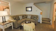 This two-story home offers a thoughtfully arranged living space designed for comfort and functionality. The main level features a cozy living room anchored by a large sectional sofa in warm, earthy tones, complemented by decorative throw pillows in floral and striped patterns. A wooden-framed mirror with ornate carved detailing adorns the wall, adding a touch of character to the neutral color palette. A low coffee table with a tray and decorative accents sits at the center, draped with a light knit throw that enhances the room's relaxed ambiance.

The living area flows naturally toward the staircase, which features light-toned wood railings and leads to the upper level, providing a clear sense of vertical space throughout the home. The carpeted flooring adds warmth underfoot, and the light-painted walls contribute to a bright, open atmosphere. The textured ceiling and understated wall décor keep the aesthetic clean and inviting. Overall, the space presents a classic, comfortable interior with a neutral design scheme well-suited to everyday living.