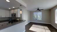This single-floor residence showcases a thoughtfully designed open-concept layout that seamlessly connects the kitchen and living areas. The kitchen is outfitted with sleek white cabinetry, dark granite countertops, and stainless steel appliances, including a built-in microwave. A stylish pendant light hangs above the kitchen bar, adding a warm, inviting accent to the space. Dark hardwood flooring runs throughout, providing a rich contrast against the neutral gray walls and crisp white trim and baseboards. The living area is bright and airy, benefiting from a large window dressed with horizontal blinds that frames a lush, green outdoor view and invites generous natural light. A ceiling fan is centrally positioned in the living area, ensuring year-round comfort. The overall ambiance is clean, contemporary, and welcoming, with a cohesive color palette of soft grays, whites, and warm wood tones that create a harmonious and functional living environment. The open floor plan enhances the sense of spaciousness, making the interior feel expansive and easy to move through.