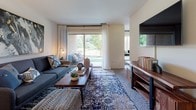 Located at 1029 Spa Rd in Annapolis, Maryland, this single-floor residence offers a thoughtfully designed layout with a cohesive coastal-inspired aesthetic throughout. The living room serves as the heart of the home, featuring large sliding glass doors that open to an outdoor patio, filling the space with natural light and views of lush greenery. The open flow leads to a defined dining area, accented by a round table, navy chairs, and decorative mirrors that add depth to the space. A wall-mounted television and a natural wood media console anchor the living area with both function and style. The galley-style kitchen is efficiently appointed with dark wood cabinetry, granite-toned countertops, hardwood flooring, and black appliances, including a full refrigerator and range. The primary bedroom is generously sized with multiple windows, soft carpeting, and direct access to a private en-suite bathroom featuring dark wood vanity cabinetry, a tub-shower combination, and hardwood-style flooring. A second bedroom carries a charming nautical theme, with decorative oars displayed on the wall. A spacious walk-in closet provides ample storage. The overall ambiance is warm and inviting, blending coastal character with practical, comfortable living in the heart of Annapolis.