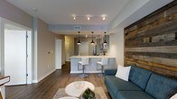 This single-floor space showcases a thoughtfully designed open-concept layout that seamlessly connects the living and kitchen areas. The kitchen is a standout feature, equipped with stainless steel appliances, a sleek white peninsula countertop, and two modern bar stools that invite casual dining. Three matte black pendant lights hang overhead, complementing the track lighting above and adding a contemporary industrial touch. Gray cabinetry and clean lines give the kitchen a polished, modern aesthetic.

The living area exudes a relaxed yet stylish ambiance, anchored by a teal sofa and a round white coffee table set atop a neutral area rug. A striking reclaimed wood accent wall adds warmth and texture, creating a focal point that balances the otherwise clean, modern palette. Rich hardwood-style flooring runs throughout, tying the spaces together cohesively.

Neutral gray and white walls are paired with recessed lighting and track fixtures to ensure the space feels bright and well-illuminated. A hallway visible from the main living area leads to additional rooms, suggesting a functional and efficient floor plan. Overall, this space combines modern design elements with warm, inviting touches to create a comfortable and visually appealing environment.