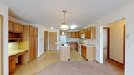 This single-floor space showcases an open-concept layout that seamlessly connects the kitchen and living areas. The kitchen is a functional centerpiece, featuring warm honey-toned wood cabinetry that extends to the ceiling, offering generous storage throughout. A central island with a built-in sink provides additional workspace and serves as a natural dividing point between the kitchen and the adjacent living area.

White appliances, including a refrigerator, electric range, and microwave, complement the light countertops and contribute to a clean, cohesive aesthetic. A pendant light hangs above the island, adding a focal point and warm illumination to the workspace, while recessed and flush-mount lighting brightens the rest of the kitchen.

The flooring transitions from resilient vinyl in the kitchen to plush carpeting in the living area, defining each zone while maintaining an open, connected feel. Textured ceilings add subtle depth throughout the space. A hallway visible in the background leads to additional rooms, suggesting a practical and well-organized floor plan. Overall, the space conveys a comfortable, functional atmosphere with a neutral palette suited to a variety of styles.