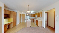 This single-floor space showcases an open-concept layout that seamlessly connects the kitchen and living areas. The kitchen is a functional centerpiece, featuring warm honey-toned wood cabinetry that extends to the ceiling, offering generous storage throughout. A central island with a built-in sink provides additional workspace and serves as a natural dividing point between the kitchen and the adjacent living area.

White appliances, including a refrigerator, electric range, and microwave, complement the light countertops and contribute to a clean, cohesive aesthetic. A pendant light hangs above the island, adding a focal point and warm illumination to the workspace, while recessed and flush-mount lighting brightens the rest of the kitchen.

The flooring transitions from resilient vinyl in the kitchen to plush carpeting in the living area, defining each zone while maintaining an open, connected feel. Textured ceilings add subtle depth throughout the space. A hallway visible in the background leads to additional rooms, suggesting a practical and well-organized floor plan. Overall, the space conveys a comfortable, functional atmosphere with a neutral palette suited to a variety of styles.