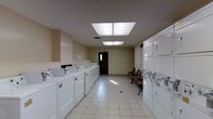 Located at 6175 Northgate Rd in Columbus, Ohio, this single-floor communal laundry facility is designed to serve residents with efficiency and convenience. The room features a generous row of top-load washing machines lining one wall, complemented by a bank of stacked dryers along the opposite side, providing ample capacity for a high volume of use. The space is finished with neutral beige walls and durable light-colored tile flooring that is easy to maintain and keeps the area feeling clean and bright. Overhead, large recessed fluorescent light panels illuminate the entire room evenly, ensuring excellent visibility throughout. A seating area with wooden chairs is positioned near the far end of the room, offering residents a comfortable place to wait. A dark-framed exit door at the rear provides convenient access, and a smoke detector is visible on the ceiling, reflecting attention to safety. The overall layout is open and well-organized, allowing for easy movement between machines. The facility presents a practical and well-maintained environment that meets the everyday laundry needs of its users.