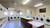 This single-story communal laundry facility is designed for both efficiency and comfort. The room is bright and airy, with light-toned walls and large windows that allow natural light to filter in, complemented by overhead fluorescent lighting and a ceiling fan that promotes air circulation throughout the space. Multiple front-loading washers and dryers are arranged along the perimeter walls, providing ample capacity for residents or guests. Centrally positioned dark wood-base folding tables with white laminate surfaces offer generous workspace for sorting and folding laundry. The flooring consists of neutral ceramic tile, which is both practical and easy to maintain. A decorative shelf near the windows adds a subtle finishing touch, adorned with small potted plants that bring a welcoming feel to the utilitarian space. An electrical panel is conveniently accessible on one wall. Overall, the facility balances functionality with a clean, well-maintained environment, making it a practical and user-friendly space for everyday laundry needs.
