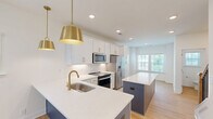Located at 2312 Ben Craig Dr in Charlotte, NC, this three-story townhome offers a well-organized layout spread across three floors, with three bedrooms, five bathrooms, and an attached garage. The ground floor houses the garage, equipped with an automatic opener and side windows for natural light, providing convenient and secure parking. The main living level features a bright, open-concept kitchen and living room combination with light hardwood-style flooring throughout. The kitchen is appointed with white shaker cabinetry, gold hardware, stainless steel appliances, a bold navy blue island with a white quartz countertop, and gold pendant lighting — creating a stylish, modern focal point. The living area benefits from multiple windows and a ceiling fan, maintaining an airy and comfortable atmosphere. A powder bathroom on this floor features a pedestal sink, oval mirror, and a window overlooking the greenery outside. The upper floors accommodate the bedrooms, each with hardwood-style flooring and ample natural light from multiple windows. The primary bedroom includes its own bathroom, featuring marble-tiled walk-in shower with a glass enclosure and a clean white vanity. Closets and a hallway provide additional storage and connectivity. The home's exterior reflects a classic board-and-batten design with a private balcony, set within a well-maintained neighborhood with lush green surroundings.