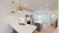 Located at 2312 Ben Craig Dr in Charlotte, NC, this three-story townhome offers a well-organized layout spread across three floors, with three bedrooms, five bathrooms, and an attached garage. The ground floor houses the garage, equipped with an automatic opener and side windows for natural light, providing convenient and secure parking. The main living level features a bright, open-concept kitchen and living room combination with light hardwood-style flooring throughout. The kitchen is appointed with white shaker cabinetry, gold hardware, stainless steel appliances, a bold navy blue island with a white quartz countertop, and gold pendant lighting — creating a stylish, modern focal point. The living area benefits from multiple windows and a ceiling fan, maintaining an airy and comfortable atmosphere. A powder bathroom on this floor features a pedestal sink, oval mirror, and a window overlooking the greenery outside. The upper floors accommodate the bedrooms, each with hardwood-style flooring and ample natural light from multiple windows. The primary bedroom includes its own bathroom, featuring marble-tiled walk-in shower with a glass enclosure and a clean white vanity. Closets and a hallway provide additional storage and connectivity. The home's exterior reflects a classic board-and-batten design with a private balcony, set within a well-maintained neighborhood with lush green surroundings.