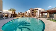 Located at 5555 W Ina Road in Marana, Arizona, this outdoor amenity space offers a resort-style experience designed for relaxation and recreation under the Sonoran Desert sky. At the heart of the space is a generously sized, freeform swimming pool with crystal-clear turquoise water, complemented by a separate spa featuring an accessible pool lift for inclusive use. The pool deck is finished with elegant paver stonework that extends throughout the entire outdoor area, providing a clean and cohesive aesthetic. Surrounding the pool, an array of lounge chairs and chaise lounges offer ample seating, while multiple shaded cabanas—draped with flowing curtains—provide private retreat areas with wicker seating and side tables. Tall, mature palm trees are strategically placed throughout the space, adding a tropical ambiance and natural shade. A well-manicured artificial turf lawn area with Adirondack chairs and patio umbrellas creates an inviting casual seating zone. The space is framed by modern multi-story residential buildings with a contemporary Southwestern architectural style, featuring stucco exteriors in warm earth tones. In the backdrop, sweeping views of the surrounding mountain ranges complete the setting, reinforcing the open, sun-drenched character of this exceptional outdoor amenity space.
