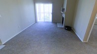 This single-floor space centers around a spacious living room finished with wall-to-wall carpeting in a neutral tone, creating a soft and cohesive foundation throughout. The room is anchored by a fireplace framed with a tiled hearth and a simple white mantle, adding a classic architectural focal point. A sliding glass door fitted with vertical blinds fills the space with natural light and provides direct access to an outdoor balcony, where greenery is visible beyond the railing. The neutral-toned walls complement the light-colored trim and baseboards, contributing to a clean and understated aesthetic. The open layout of the living area allows for flexible furniture arrangements, making it adaptable to various needs. The combination of the fireplace, natural light, and outdoor access gives the space a balanced character that is both practical and inviting. The tiled transition near the fireplace hearth provides a subtle contrast to the surrounding carpet, adding a minor design detail to the room's overall composition.