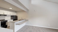 This single-floor residence offers a clean, functional layout with a neutral color palette throughout. The kitchen is a standout feature, designed with white shaker-style cabinetry providing ample storage, light speckled countertops, and a peninsula that doubles as a breakfast bar. Black appliances — including a gas range and refrigerator — add a bold contrast to the bright, white cabinetry. Wood-look vinyl flooring runs through the kitchen, lending a warm, easy-to-maintain surface underfoot.

The living area transitions seamlessly from the kitchen peninsula and is characterized by impressively vaulted ceilings that lend a sense of openness and volume to the space. Soft, neutral carpet continues into the bedroom, which benefits from a generously sized window that invites natural light and frames views of surrounding greenery. The vaulted ceiling in the bedroom further enhances the airy feel of the room.

The bathroom is neatly appointed with wood-look vinyl flooring, a tiled tub and shower combination, a toilet, and a vanity topped with a speckled countertop and an integrated mirror. Clean white finishes throughout maintain a cohesive, bright aesthetic. Overall, the residence presents a well-organized and comfortable living environment with practical finishes and thoughtful design details.