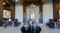 This single-story space presents a sophisticated entryway that sets a refined tone throughout. The foyer is defined by soaring ceiling height with a bold, dark-toned upper border that frames the space and draws the eye upward, creating a dramatic architectural statement. The walls feature a two-tone treatment, with warm charcoal-brown paneling flanking a central recessed alcove that leads to a set of classic French doors. The doors, fitted with divided-light glass panels, allow natural light to filter into the entry and provide a glimpse of the exterior beyond. Light-toned tile flooring reflects the ambient light, enhancing the overall brightness of the space. A round, high-gloss black accent table anchors the center of the foyer, displaying a curated collection of textured metallic vases in silver tones that serve as a focal decorative element. To one side, a richly upholstered chair and a patterned footstool introduce a hint of color and comfort. Recessed ceiling lighting supplements the natural light, ensuring the space remains well-illuminated. Overall, the entryway balances bold architectural details with carefully chosen furnishings to create a polished and welcoming first impression.