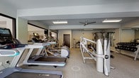 This modern fitness center is thoughtfully designed to support a full range of workout routines in a spacious, single-floor layout. The space is divided into distinct zones, seamlessly connected to create a cohesive training environment. The cardio area is outfitted with multiple treadmills, elliptical machines, and stationary bikes, all positioned to take advantage of large windows that invite natural light and offer views of the surrounding building exterior. Ceiling fans are installed throughout, ensuring consistent air circulation across the entire facility. The strength training zone features a robust selection of equipment, including cable machines, multi-function weight stations, adjustable benches, and a well-stocked dumbbell rack spanning a range of weights. Full-length mirrors line key sections of the walls, enhancing the sense of openness while allowing users to monitor their form during workouts. The flooring throughout is a durable, neutral-toned carpet well-suited for a fitness environment. Recessed and flush-mounted fluorescent lighting provides consistent, bright illumination across the entire space. A private room with a dark-finished door offers an additional enclosed area within the facility. The overall ambiance is clean, contemporary, and functional, making this fitness center a well-rounded amenity for residents or members seeking a professional-grade workout experience.