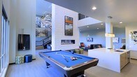 This dynamic amenity lounge is designed with a contemporary aesthetic and an open, airy layout that encourages both active recreation and relaxed socializing. At the forefront of the space sits a full-sized pool table with a vibrant blue felt surface, serving as a natural centerpiece for the room. Behind it, a striking white brick column anchors a sleek, linear gas fireplace, above which a framed artwork adds a decorative focal point. The soaring ceiling height lends the space a dramatic, expansive feel, amplified by recessed lighting and a sculptural spiral pendant fixture that draws the eye upward. A lounge area is arranged nearby, furnished with modern seating in neutral tones accented by pops of orange, creating an inviting spot to unwind. A wall-mounted television adds to the entertainment-oriented atmosphere. In the background, a staircase with a sleek metal railing leads to an upper level, and large-scale artwork decorates the adjacent wall, adding visual depth and personality. The light hardwood flooring and cool blue-toned lighting throughout unify the space with a cohesive, modern ambiance.