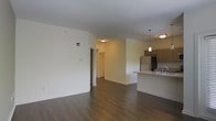 This single-floor space showcases an open-concept design that seamlessly connects the living area and kitchen. Dark hardwood flooring extends throughout, providing a warm contrast against the crisp white walls and white baseboards. The living area is generously sized, offering a blank canvas with clean lines and a neutral palette.

The kitchen is positioned along one side of the space and features a raised breakfast bar countertop, medium-toned wood cabinetry, and stainless steel appliances including a refrigerator. Pendant lighting fixtures hang above the counter, adding functional illumination and a subtle decorative touch. A sink is integrated into the countertop, and additional cabinetry lines the wall for ample storage.

A hallway visible from the main living area leads to additional spaces within the unit, hinting at further rooms beyond the open zone. Ceiling-mounted lighting provides consistent illumination throughout. The overall ambiance is modern and understated, with a practical, well-organized layout suited for everyday living. The open flow between the kitchen and living areas creates a spacious, connected feel.