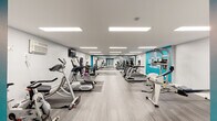 This fitness center is a comprehensively outfitted facility designed to support a full range of workout routines. The space features a clean, modern aesthetic with crisp white walls accented by bold teal detailing and a motivational wall graphic that reads "The Only Bad Workout Is The One You Didn't Do," reinforcing an energizing atmosphere throughout.

The main workout floor is finished with wood-look luxury vinyl plank flooring and padded gym mats strategically placed beneath equipment. Recessed panel lighting runs across the white ceiling, ensuring the space is brightly and evenly lit. The layout is open and spacious, allowing for comfortable movement between stations.

Cardio equipment includes several treadmills, elliptical trainers, stationary bikes, and a rowing machine, all well-spaced across the floor. Strength training options include a multi-function cable machine, a leg press unit, and a dumbbell rack stocked with a full range of free weights. A large wall mirror spans one end of the room, visually expanding the space and allowing users to monitor their form.

Additional amenities include a flat-screen television mounted on the wall, a water fountain, storage lockers, and a wall-mounted AC unit. A dedicated open mat area provides room for stretching, core work, or floor-based exercises. The overall environment is clean, organized, and purpose-built for an effective fitness experience.