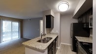 This single-floor space showcases a thoughtfully designed open-concept layout where the kitchen connects seamlessly to the living area. The kitchen is a standout feature, equipped with dark espresso-toned cabinetry that contrasts sharply against light granite countertops with subtle veining. A double stainless steel sink is set into the peninsula, which also functions as a breakfast bar, offering additional seating and separating the kitchen from the adjoining living space. Stainless steel appliances, including a full-size refrigerator, complete the modern kitchen setup. Upper cabinetry provides generous storage, while lower cabinets offer additional organization options. The flooring throughout features light-toned wood-look planks that add warmth to the space. Walls are painted in a cool, neutral gray tone, contributing to a cohesive and contemporary aesthetic. A textured ceiling adds subtle dimension overhead, and flush-mount lighting ensures the kitchen area is well illuminated. The open sightline into the living area enhances the overall sense of space, making the layout feel connected and functional throughout.