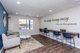 Located at 10100 Brown Bridge Rd in Covington, Georgia, this single-floor professional office space belongs to Patrician Management and is thoughtfully designed to create a welcoming, polished environment for clients and staff alike. Rich hardwood-style flooring flows throughout, tying together each area with a cohesive and modern aesthetic.

The main common area serves as both a reception and lounge space, featuring a wall-mounted TV, a beverage station, and a high-top bar counter with seating — ideal for informal meetings or client check-ins. A bold wall decal reading "We make living easy. – Patrician Management" anchors the space with the company's brand identity.

The waiting area is warmly appointed with neutral upholstered seating, a decorative area rug, and a marble-top coffee table. A striking teal accent wall paired with a round gold mirror and large windows flooding the room with natural light creates a calm, inviting atmosphere.

A private consultation nook features a vibrant yellow patterned armchair, a wooden side table with fresh floral arrangement, colorful artwork, and bold navy hexagonal acoustic panels — offering a creative and comfortable setting for one-on-one interactions.

The private office is equipped with a rustic wood desk, ergonomic seating, a tiered corner shelving unit with décor, and a decorative wooden U.S. map — providing a functional yet personable workspace. Overall, the space balances professionalism with warmth.