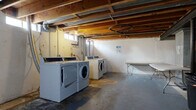 This basement-level space is designed to serve practical utility and storage needs. The layout is divided into two primary zones: a shared laundry area and an open storage section. The laundry area is equipped with multiple commercial-grade washers and dryers arranged side by side, providing ample capacity for shared use. A folding table is also present, offering a convenient surface for laundry tasks. Overhead, exposed wood joists, ventilation ducts, and utility piping run along the unfinished ceiling, giving the space a raw, functional character. Small hopper windows along the upper portion of the walls allow limited natural light to filter in. The flooring throughout is bare concrete, consistent with the utilitarian nature of the space. The storage section features an expansive open floor plan supported by structural steel columns with black painted finishes. Along one side, enclosed chain-link storage units provide secure, individual storage compartments. The white-painted concrete block walls and overhead artificial lighting illuminate the area adequately. The mechanical systems, including HVAC equipment, are visible and accessible within the space. Overall, this basement offers a straightforward, no-frills environment purpose-built for shared laundry and storage functions.