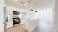 This thoughtfully designed single-floor residence offers a cohesive, modern layout across its one-bedroom, one-bathroom floor plan. The kitchen serves as a focal point, featuring crisp white shaker cabinetry, a white subway tile backsplash, a full suite of stainless steel appliances including a refrigerator, range, microwave, and dishwasher, and a central island topped with a light quartz countertop. Two statement gold pendant lights hang above the island, adding a warm, contemporary accent. Light wood-look flooring extends throughout the kitchen and living area, enhancing the bright, airy atmosphere created by recessed lighting and a ceiling fan. The open layout allows the kitchen to flow naturally into the living space, which benefits from natural light through a window. The bedroom is carpeted for comfort and includes a ceiling fan, a window with blinds, and access to the full bathroom. The bathroom is elongated and well-appointed, with a white vanity offering ample drawer storage, a large mirror, a bathtub with shower fixtures, and wood-look flooring. Directly accessible from the bathroom is a dedicated walk-in closet, carpeted and fitted with multiple hanging rods and shelving, providing generous storage for everyday needs.