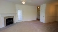 This single-floor space centers around a spacious living area finished with wall-to-wall beige carpeting that lends a warm, neutral foundation throughout. The room features a fireplace with a classic white mantel positioned along one wall, adding a functional and visually grounding element to the space. A ceiling fan with a built-in light fixture is mounted overhead, providing both air circulation and ambient lighting.

Natural light enters through a white-framed exterior door fitted with a window and blinds, offering a practical connection to the outdoors while brightening the interior. The walls are finished in a clean, off-white tone that complements the light carpeting and contributes to an airy, open feel.

Two interior doorways are visible from the main living area. One leads to what appears to be a hallway or additional room illuminated by a ceiling-mounted light, while the other provides access to a closet or secondary space. The overall layout suggests a straightforward, functional floor plan with a cohesive and understated aesthetic throughout.