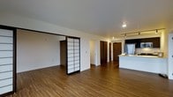 This single-floor space blends modern design with subtle Japanese-inspired elements, creating a clean and functional living environment. The open-concept layout allows for a seamless flow between the main living area and the kitchen, giving the space an airy, expansive feel. Warm-toned hardwood flooring runs throughout, adding a natural richness to the interior and grounding the overall aesthetic.

A standout feature of the space is the traditional shoji-style sliding door, framed in dark metal with translucent panels. This architectural detail introduces a refined, cultural design element that adds visual interest and serves as a flexible room divider. The white walls and smooth ceilings keep the atmosphere bright and uncluttered.

The kitchen is positioned toward the rear of the open floor plan and features stainless steel appliances, a light-colored countertop peninsula, and track lighting overhead — providing both task illumination and a modern finishing touch. Dark-toned interior doors lead to additional rooms, contributing to a cohesive and intentional color palette throughout. Recessed lighting complements the track fixtures, ensuring the space is well-lit from multiple sources. Overall, the space offers a thoughtful combination of contemporary design and understated cultural character.