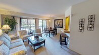 This single-floor residence offers a thoughtfully designed layout that balances comfort and functionality across its key living spaces. The open-plan living and dining area is a standout feature, bathed in natural light streaming through large sliding glass doors that lead to a private deck overlooking lush, mature greenery. A classic chandelier anchors the dining space, which comfortably accommodates a full dining set, while the adjoining living room provides a relaxed atmosphere with soft carpeting and neutral-toned walls. A pass-through window connects the kitchen to the living area, enhancing the sense of openness. The kitchen is equipped with ample white cabinetry, generous counter space, a built-in stovetop, and a dishwasher, offering practical utility in a compact footprint. The primary bedroom features plush carpeting, a built-in closet with bifold doors, and dedicated space for a full dresser suite. The second bedroom is equally comfortable, with a window allowing natural light and sufficient space for a full bed and additional furnishings. The bathroom is finished with classic white subway tile in the tub-shower surround, a single vanity, and a well-lit mirror. Throughout the residence, neutral tones and carpet flooring create a cohesive, understated aesthetic.