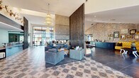 This single-floor community space blends contemporary design with eclectic character, creating a welcoming and visually rich environment throughout. The main lounge area immediately impresses with soaring ceilings, ornate tiered crystal chandeliers, and intricate patterned tile flooring that anchors the room with a bold, artistic flair. A striking double-sided concrete fireplace serves as the centerpiece, flanked by a tall vertical wood slat partition that elegantly divides the space while maintaining an open, airy flow. Plush blue-grey seating arrangements invite comfortable gathering, while exposed brick accent walls add warmth and texture. Floor-to-ceiling steel-framed glass windows and doors flood the interior with abundant natural light and offer sweeping views of the surrounding outdoor areas. A dedicated billiards room features a handsome wooden pool table set beneath a sculptural branching chandelier with globe pendants, complemented by exposed brick, large windows, and intimate bistro seating. The reception area showcases a dramatic black marble counter with a warm wood base, recessed lighting, and French doors leading to adjacent rooms. Decorative antler mounts and curated artwork throughout contribute to the space's distinctive, lodge-inspired personality, making it both functional and memorable.
