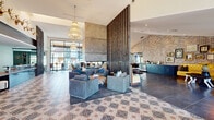This single-floor community space blends contemporary design with eclectic character, creating a welcoming and visually rich environment throughout. The main lounge area immediately impresses with soaring ceilings, ornate tiered crystal chandeliers, and intricate patterned tile flooring that anchors the room with a bold, artistic flair. A striking double-sided concrete fireplace serves as the centerpiece, flanked by a tall vertical wood slat partition that elegantly divides the space while maintaining an open, airy flow. Plush blue-grey seating arrangements invite comfortable gathering, while exposed brick accent walls add warmth and texture. Floor-to-ceiling steel-framed glass windows and doors flood the interior with abundant natural light and offer sweeping views of the surrounding outdoor areas. A dedicated billiards room features a handsome wooden pool table set beneath a sculptural branching chandelier with globe pendants, complemented by exposed brick, large windows, and intimate bistro seating. The reception area showcases a dramatic black marble counter with a warm wood base, recessed lighting, and French doors leading to adjacent rooms. Decorative antler mounts and curated artwork throughout contribute to the space's distinctive, lodge-inspired personality, making it both functional and memorable.