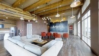 This single-floor space exudes an industrial-chic aesthetic, blending raw architectural elements with modern furnishings to create an inviting and dynamic communal environment. Exposed brick walls add warmth and texture throughout, while rustic wooden ceiling beams lend a sense of heritage and craftsmanship. The ceiling is adorned with an eclectic mix of pendant lights — from clustered Edison bulbs to broad industrial-style shades — casting a warm, layered glow across the room. The flooring transitions between rich dark hardwood and lighter wood tones, subtly delineating different zones within the open layout. A generous seating area anchors the space with plush, neutral-toned sofas arranged around a reclaimed wood coffee table, ideal for relaxed gatherings. Adjacent to this, a cluster of bold terracotta-red lounge chairs surrounds low tables, offering a more intimate setting. A striking large-scale cityscape mural rendered in metallic tones on a dark background serves as a dramatic focal point on one wall, adding an urban artistic flair. The open floor plan and generous ceiling height contribute to an airy, loft-like atmosphere, making this space well-suited for socializing, lounging, or community events.