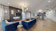This well-appointed single-floor amenity space is designed to serve as a sophisticated social hub, combining comfort and modern design throughout its open, flowing layout. The main lounge area serves as the centerpiece, anchored by a sleek linear fireplace set against a floor-to-ceiling dark tile accent wall. Plush navy velvet sofas, rust-toned ottomans, and a striking Sputnik chandelier lend the space a curated, upscale aesthetic, while warm hardwood-style flooring ties the entire area together. Large windows dressed with sheer white curtains bathe the room in natural light and offer views to the outdoors, enhancing the open and airy ambiance. Adjacent to the lounge, a dining zone provides additional seating with mid-century modern chairs around a round table, ideal for casual gatherings. The kitchen is fully equipped with stainless steel appliances, a range, dishwasher, light wood cabinetry, tile backsplash, and ample counter space, complemented by a large granite-topped island with bar-height seating and decorative pendant lighting. A hallway corridor features glass-partitioned rooms with wide-plank flooring and large-format tile, providing flexible spaces suitable for meetings or private use. Colorful artwork adorns the walls, adding personality and vibrancy throughout.