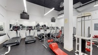 This fitness center is a dedicated single-floor workout facility designed to meet a wide range of exercise needs. The space features a dark drop ceiling with exposed industrial elements, contrasted by bright fluorescent strip lighting that ensures a well-lit environment throughout. White structural columns and clean white walls contribute to an open, airy feel, while large wall-mounted mirrors along one side visually expand the space and allow users to monitor their form during workouts.

The flooring consists of durable dark rubber matting, ideal for heavy equipment use and providing a non-slip surface for safety. The cardio section is well-stocked with treadmills, elliptical machines, and stair climbers, all equipped with individual digital consoles. Strength training options include a red-upholstered weight bench and a multi-use cable machine positioned along the perimeter. Multiple wall-mounted flat-screen televisions are strategically placed throughout, providing entertainment during workouts.

The layout is thoughtfully organized to maximize floor space and allow comfortable movement between stations. The overall ambiance is functional and professional, making it a practical and fully equipped environment for a focused fitness routine.