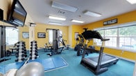 This fitness center is designed to support a comprehensive workout experience within a single, well-organized space. The room is immediately distinguished by its bold yellow walls, which create an energizing and motivating atmosphere. Bright flush-mounted ceiling panels provide consistent, even lighting throughout the area, ensuring excellent visibility for all activities.

The space is outfitted with a diverse range of exercise equipment. Cardio options include a NordicTrack treadmill and an elliptical machine, both positioned near windows that allow natural light to enter and offer views of the surrounding greenery. For strength training, a multi-station cable and weight machine anchors the center of the room, complemented by a full rack of dumbbells along the wall for free-weight exercises. A stability ball and a foldable exercise mat are also available, catering to stretching, core work, and floor-based routines.

The flooring is covered in a teal-toned carpet, adding a layer of comfort underfoot while contributing to the room's vibrant color palette. Motivational artwork and informational posters accent the walls, reinforcing the space's purpose. Windows on multiple walls promote natural ventilation and a connection to the outdoors. Overall, the fitness center is practical, inviting, and thoughtfully arranged to accommodate a variety of training needs.