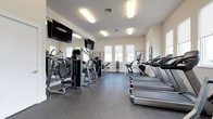 This fitness center is a comprehensive, single-floor facility designed to support a full range of workout routines. The space is bright and open, with multiple windows fitted with roller shades that allow natural light to fill the room while providing privacy when needed. The durable speckled rubber flooring throughout offers both safety and comfort underfoot, making it well-suited for high-intensity exercise.

The cardio area is equipped with a row of treadmills and elliptical machines, providing ample options for aerobic training. A dedicated strength training section features a variety of cable and resistance machines, a rowing machine, a weight bench, and a dumbbell rack with a graduated selection of free weights. Full-length wall mirrors run along one side of the space, enhancing the sense of openness and allowing users to monitor their form during workouts.

Entertainment is provided by multiple large flat-screen TVs mounted on the walls, ensuring an engaging environment during exercise sessions. Athletic-themed framed artwork decorates the walls, contributing to a motivating atmosphere. The facility is accessed through a set of glass double doors and is well-lit by recessed fluorescent ceiling fixtures. The clean, neutral color palette and organized layout create a functional, welcoming environment suited to a variety of fitness levels and goals.