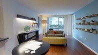 This well-appointed single-floor residence offers a thoughtfully designed living area that balances comfort and contemporary style. The space is anchored by rich hardwood flooring that runs throughout, lending warmth and a sense of cohesion to the interior.

The living area is the focal point of the home, centered around a plush taupe sofa accented with a vibrant orange throw pillow and complemented by a distinctive yellow tufted oval ottoman, adding a pop of color and personality to the space. A large picture window dominates one wall, inviting abundant natural light and offering a pleasant view of lush greenery outside, creating a seamless connection between the interior and the surrounding landscape. Sheer white curtains frame the window elegantly, softening the light that fills the room.

Decorative details add character throughout, including a sleek console table with understated décor and illuminated display shelving that lines one wall, offering both functional storage and an artistic touch. A round dark-toned dining or accent table in the foreground adds contrast and visual interest. The walls are finished in a clean white palette with a cool blue accent, contributing to the bright and airy ambiance of the overall space.