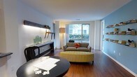 This well-appointed single-floor residence offers a thoughtfully designed living area that balances comfort and contemporary style. The space is anchored by rich hardwood flooring that runs throughout, lending warmth and a sense of cohesion to the interior.

The living area is the focal point of the home, centered around a plush taupe sofa accented with a vibrant orange throw pillow and complemented by a distinctive yellow tufted oval ottoman, adding a pop of color and personality to the space. A large picture window dominates one wall, inviting abundant natural light and offering a pleasant view of lush greenery outside, creating a seamless connection between the interior and the surrounding landscape. Sheer white curtains frame the window elegantly, softening the light that fills the room.

Decorative details add character throughout, including a sleek console table with understated décor and illuminated display shelving that lines one wall, offering both functional storage and an artistic touch. A round dark-toned dining or accent table in the foreground adds contrast and visual interest. The walls are finished in a clean white palette with a cool blue accent, contributing to the bright and airy ambiance of the overall space.