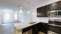 This single-floor space showcases a thoughtfully designed open-concept layout that seamlessly connects the kitchen and living areas. The kitchen serves as a central highlight, featuring rich, dark espresso-toned cabinetry that provides a striking contrast against the crisp white walls and light granite countertops. A kitchen island extends the counter space and doubles as a casual gathering point, complete with a stainless steel double-basin sink and a sleek pull-down faucet. A stainless steel dishwasher is integrated beneath the counter, adding functional convenience. Three modern cylindrical pendant lights hang above the island, providing focused task lighting while enhancing the contemporary aesthetic. The upper cabinetry offers generous storage and is accented with brushed metal hardware that complements the overall design. Light wood-style flooring runs throughout, adding warmth to the space. The adjoining living area benefits from natural light streaming through a window fitted with horizontal blinds, and a ceiling fan ensures comfortable airflow. The open sightlines between the kitchen and living space create an airy, cohesive environment ideal for both everyday living and entertaining.