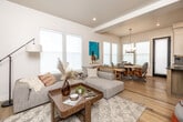 This two-story home presents a thoughtfully designed layout across two levels, combining modern finishes with a warm, approachable aesthetic throughout. The main floor centers on an open-concept living, dining, and kitchen area unified by light hardwood flooring that flows seamlessly from room to room. Large windows with roller shades flood the space with abundant natural light, enhancing the home's airy, neutral palette. A black-framed entry door adds a sharp contrast to the crisp white walls and trim.

The kitchen is a standout feature, equipped with a substantial island topped with a gray quartz countertop, warm natural wood cabinetry, two-tone upper cabinetry in white and charcoal, stainless steel appliances, and globe pendant lighting overhead. The adjacent dining area accommodates a round natural wood table beneath a stylish multi-arm chandelier, complemented by woven rattan seating.

The living area provides a comfortable, well-lit retreat with direct access to an entry nook that houses a media setup near the front door. Upstairs, the bedrooms feature plush carpet flooring, recessed lighting, and ample windows that invite natural light. The bathroom is finished with marble-look tile surrounding a soaking tub, natural wood vanity cabinetry, matte black hardware, and gray quartz countertops — delivering a clean, contemporary feel.