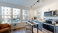This single-floor space presents a thoughtfully designed layout that balances modern functionality with distinctive decorative touches. The kitchen is a standout feature, showcasing two-tone cabinetry with white upper cabinets and slate-blue lower cabinets, complemented by stainless steel appliances including a range, microwave, and refrigerator. A matte black faucet adds a contemporary accent at the sink, while track lighting illuminates the space. A movable island with bar stools serves as both a prep and dining area. Large windows allow natural light to pour in, offering views of the surrounding urban exterior.

The living area continues the bright, airy aesthetic with white walls, light wood-style flooring, and a generous window that further connects the interior to the outside. The bedroom features a bold Art Deco-inspired gold patterned accent wall that adds warmth and personality, paired with light wood flooring.

The bathroom is clean and crisp, with white subway tile surrounding the tub-shower combo, matte black fixtures, and a white vanity. A dedicated nook with geometric-patterned wallpaper houses a compact ladder desk and shelving, providing a clever workspace solution. Throughout, the space maintains a cohesive modern ambiance.