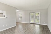 This single-story unit is thoughtfully laid out across a comfortable footprint, accommodating four bedrooms and four bathrooms connected by three hallways. The open living room serves as the central gathering space, finished with light gray wood-style laminate flooring that runs throughout the unit and a textured ceiling. French doors at the far end of the living room invite natural light and provide outdoor access, while an adjacent window further brightens the space. A pass-through window connects the living area to the kitchen, enhancing the open feel. The kitchen is well-appointed with white shaker-style cabinetry, marble-look quartz countertops, stainless steel appliances including a refrigerator, range, and microwave, and a single-basin sink. Each bedroom features neutral gray walls, windows with blinds for natural light, and closet access. The primary bedroom offers a generous layout with ample room for furnishings. The bathrooms maintain a clean, consistent aesthetic, with gray vanity cabinetry, white countertops, lighted mirrors, and white tile tub-shower surrounds with chrome fixtures throughout.