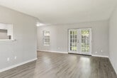 This single-story unit is thoughtfully laid out across a comfortable footprint, accommodating four bedrooms and four bathrooms connected by three hallways. The open living room serves as the central gathering space, finished with light gray wood-style laminate flooring that runs throughout the unit and a textured ceiling. French doors at the far end of the living room invite natural light and provide outdoor access, while an adjacent window further brightens the space. A pass-through window connects the living area to the kitchen, enhancing the open feel. The kitchen is well-appointed with white shaker-style cabinetry, marble-look quartz countertops, stainless steel appliances including a refrigerator, range, and microwave, and a single-basin sink. Each bedroom features neutral gray walls, windows with blinds for natural light, and closet access. The primary bedroom offers a generous layout with ample room for furnishings. The bathrooms maintain a clean, consistent aesthetic, with gray vanity cabinetry, white countertops, lighted mirrors, and white tile tub-shower surrounds with chrome fixtures throughout.