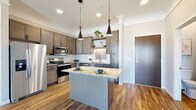 This well-designed single-floor residence offers a thoughtful layout that balances modern aesthetics with everyday functionality. The kitchen serves as a standout centerpiece, showcasing warm grey-toned cabinetry with sleek hardware, granite countertops, and a full suite of stainless steel appliances including an electric range, built-in microwave, and dishwasher. A herringbone-pattern tile backsplash adds a refined touch, while open floating shelves provide both storage and display space. A central island with a matching granite surface and pendant lighting overhead makes the kitchen ideal for casual dining and meal preparation. Rich wood-look flooring flows throughout the main living areas, anchoring the open and airy living room, which benefits from natural light streaming in through large windows and a sliding glass door that leads to a private balcony. The bedroom features plush carpeting and a bright window with neighborhood views. The bathroom is finished with the same quality cabinetry and granite countertop found in the kitchen, a single-sink vanity, and a walk-in shower. Neutral wall tones and consistent finishes throughout create a cohesive, inviting atmosphere that is both stylish and livable.
