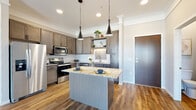 This well-designed single-floor residence offers a thoughtful layout that balances modern aesthetics with everyday functionality. The kitchen serves as a standout centerpiece, showcasing warm grey-toned cabinetry with sleek hardware, granite countertops, and a full suite of stainless steel appliances including an electric range, built-in microwave, and dishwasher. A herringbone-pattern tile backsplash adds a refined touch, while open floating shelves provide both storage and display space. A central island with a matching granite surface and pendant lighting overhead makes the kitchen ideal for casual dining and meal preparation. Rich wood-look flooring flows throughout the main living areas, anchoring the open and airy living room, which benefits from natural light streaming in through large windows and a sliding glass door that leads to a private balcony. The bedroom features plush carpeting and a bright window with neighborhood views. The bathroom is finished with the same quality cabinetry and granite countertop found in the kitchen, a single-sink vanity, and a walk-in shower. Neutral wall tones and consistent finishes throughout create a cohesive, inviting atmosphere that is both stylish and livable.