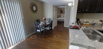 This single-floor unit offers an open-concept layout that combines the kitchen, living, and sleeping areas into one cohesive, functional space. The interior is finished with warm-toned walls and rich dark hardwood flooring that runs throughout, lending a sense of warmth and continuity to the space.

The kitchen is positioned along one side and features dark cabinetry, a stainless steel refrigerator, and a granite-topped breakfast bar that provides a natural visual divider between the cooking and living zones without sacrificing the open feel. Flush-mount ceiling lighting keeps the space well-lit throughout.

The living and sleeping area is open and flexible, currently furnished with a white daybed with built-in storage drawers beneath, making efficient use of the available space. Decorative wall art adds personality to the neutral backdrop, with pieces referencing iconic imagery. The white trim and baseboards provide a clean contrast against the darker flooring and wall tones.

A doorway leads to an adjacent room, suggesting additional utility space such as a bathroom or closet. Overall, the unit presents a practical and well-organized layout ideal for those seeking an efficiently designed, single-level living space with a warm, inviting ambiance.