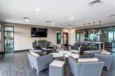 This contemporary amenity lounge is thoughtfully designed to serve as a versatile communal gathering space. The open floor plan is anchored by dark hardwood-style flooring that extends throughout, complementing the neutral warm-toned walls and creating a cohesive, modern aesthetic.

The main lounge area features multiple clusters of upholstered armchairs in gray and dark brown tones, arranged around low round tables and ottomans to encourage both casual socializing and relaxed lounging. A large wall-mounted flat-screen television is positioned above a sleek credenza with decorative greenery, serving as a focal point for the seating arrangement.

Toward one side of the space, a bar-height communal table paired with contemporary stools provides an additional area for dining or collaborative work, illuminated by a row of elegant pendant lights that add a refined touch overhead. Adjacent glass-panel partitions offer views into what appears to be a fitness area.

A standout feature of the lounge is the expansive wall of floor-to-ceiling glass doors on one side, which flood the interior with natural light and open directly onto an outdoor terrace, offering broad exterior views. Recessed ceiling lighting supplements the natural light throughout, ensuring a well-lit, welcoming atmosphere. Built-in cabinetry along one wall adds practical storage.