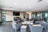 This contemporary amenity lounge is thoughtfully designed to serve as a versatile communal gathering space. The open floor plan is anchored by dark hardwood-style flooring that extends throughout, complementing the neutral warm-toned walls and creating a cohesive, modern aesthetic.

The main lounge area features multiple clusters of upholstered armchairs in gray and dark brown tones, arranged around low round tables and ottomans to encourage both casual socializing and relaxed lounging. A large wall-mounted flat-screen television is positioned above a sleek credenza with decorative greenery, serving as a focal point for the seating arrangement.

Toward one side of the space, a bar-height communal table paired with contemporary stools provides an additional area for dining or collaborative work, illuminated by a row of elegant pendant lights that add a refined touch overhead. Adjacent glass-panel partitions offer views into what appears to be a fitness area.

A standout feature of the lounge is the expansive wall of floor-to-ceiling glass doors on one side, which flood the interior with natural light and open directly onto an outdoor terrace, offering broad exterior views. Recessed ceiling lighting supplements the natural light throughout, ensuring a well-lit, welcoming atmosphere. Built-in cabinetry along one wall adds practical storage.