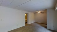 This single-floor space presents a straightforward and functional layout centered around a generously sized main room. The room is finished with wall-to-wall beige carpeting that adds a layer of warmth underfoot, while the white walls and flat white ceiling contribute to a clean, neutral aesthetic throughout. A ceiling fan with an integrated light fixture is centrally mounted, providing both air circulation and ambient lighting to the space.

The room's open layout allows for flexible furniture arrangements, making it adaptable to a variety of uses. An interior doorway leads directly to an adjoining bathroom, visible from the main room and illuminated by warm lighting. The bathroom appears to include a vanity with a sink, offering basic but practical amenities.

The overall ambiance of the space is understated and utilitarian, characterized by its simple design elements and muted color palette. The absence of windows visible in this view gives the room a more enclosed feel, though the light-colored walls and ceiling help maintain a sense of openness. The space is well-suited for those seeking a no-frills, move-in-ready environment with a clean and uncluttered foundation.